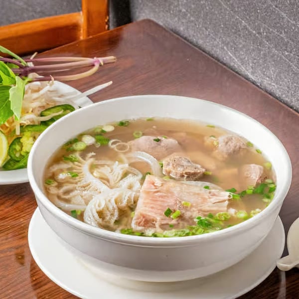 A bowl of Vietnamese pho with noodles, meat, and green onions, served with a side of fresh herbs.