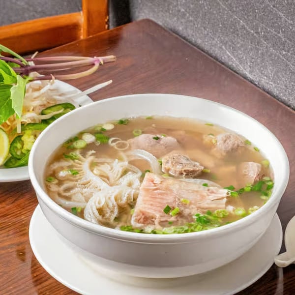 A bowl of Vietnamese pho with noodles, meat, and green onions, served with a side of fresh herbs.